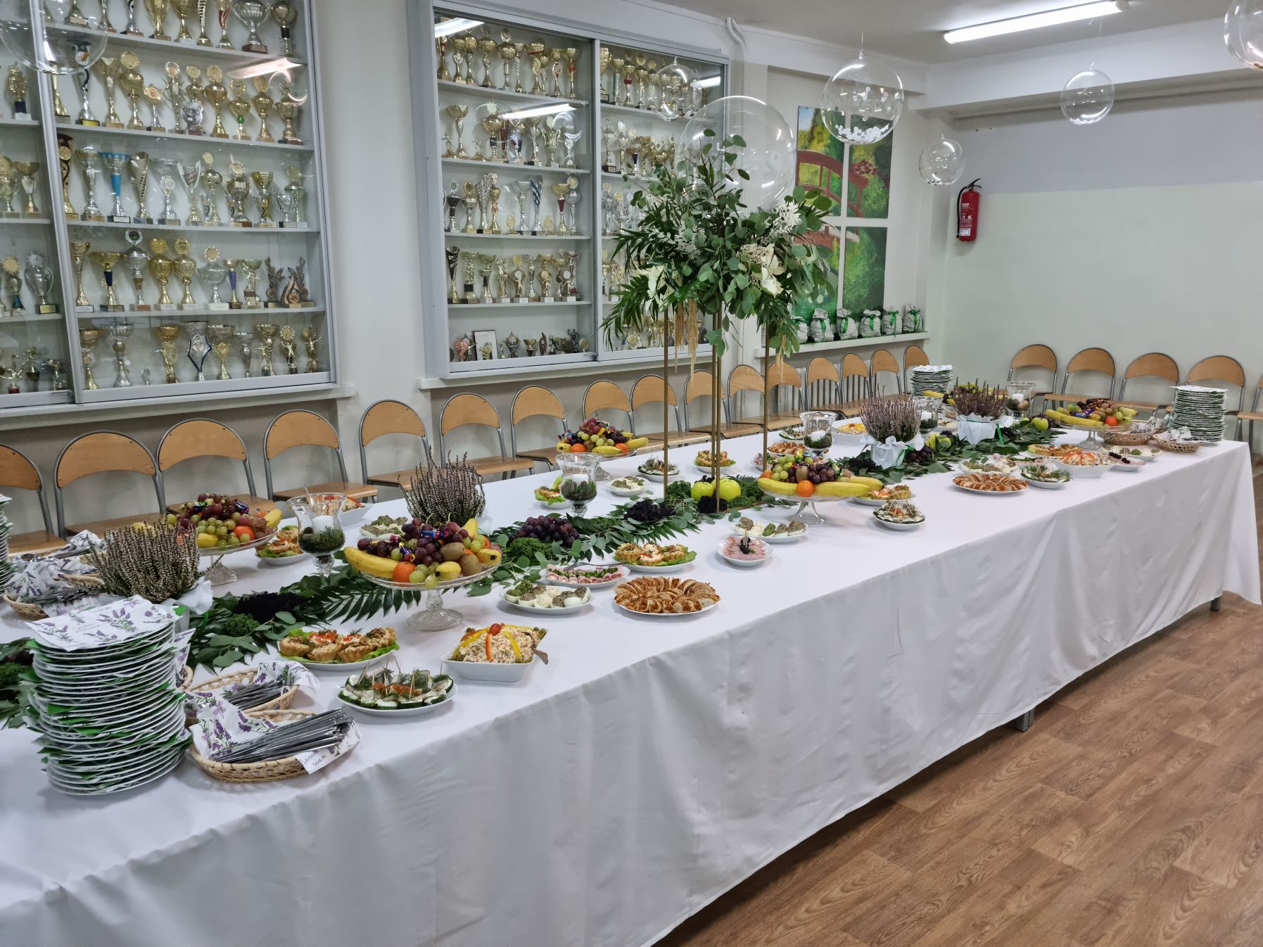 Długi stół z białym obrusem, na stole potrawy i dekoracje kwiatowe
