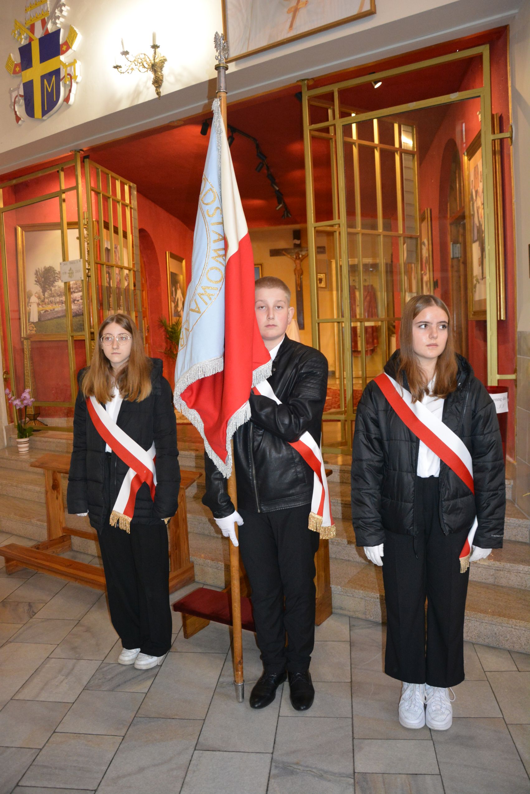 Wnętrze kościoła, poczet sztandarowy, w środku chłopiec trzyma sztandar, po obu stronach dziewczynki, wszyscy przepasani biało-czerwoną szarfą