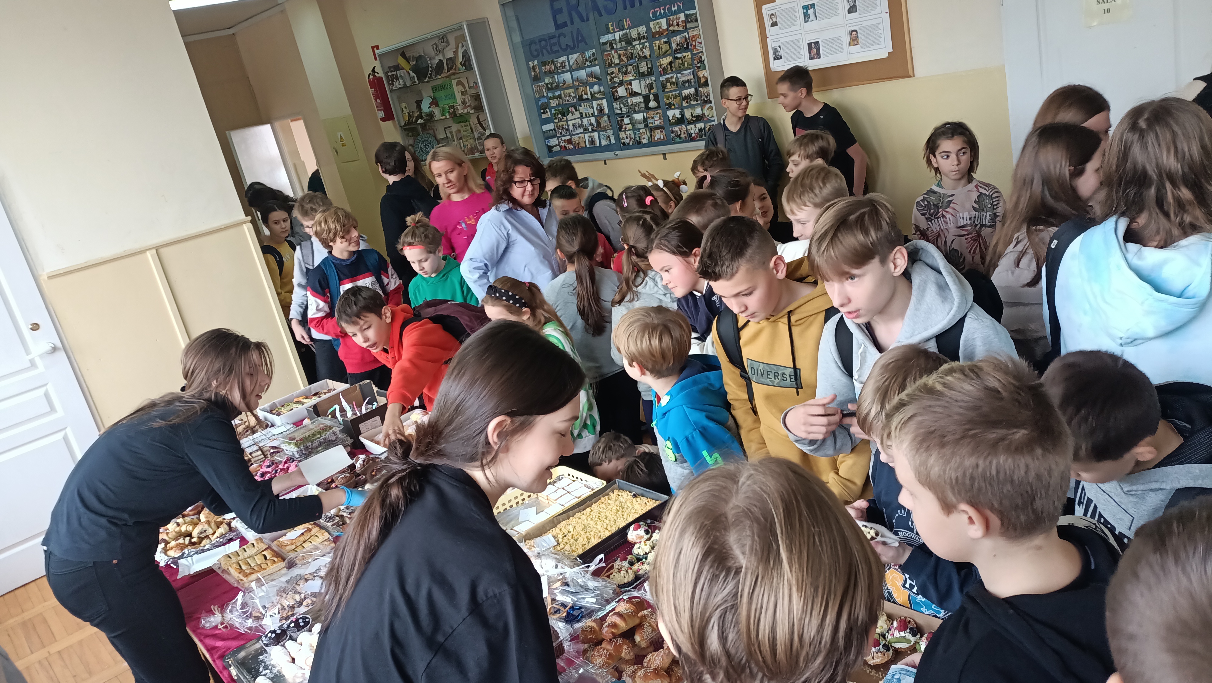 Uczniowie i nauczyciele stoją przy stołach i kupują słodkości