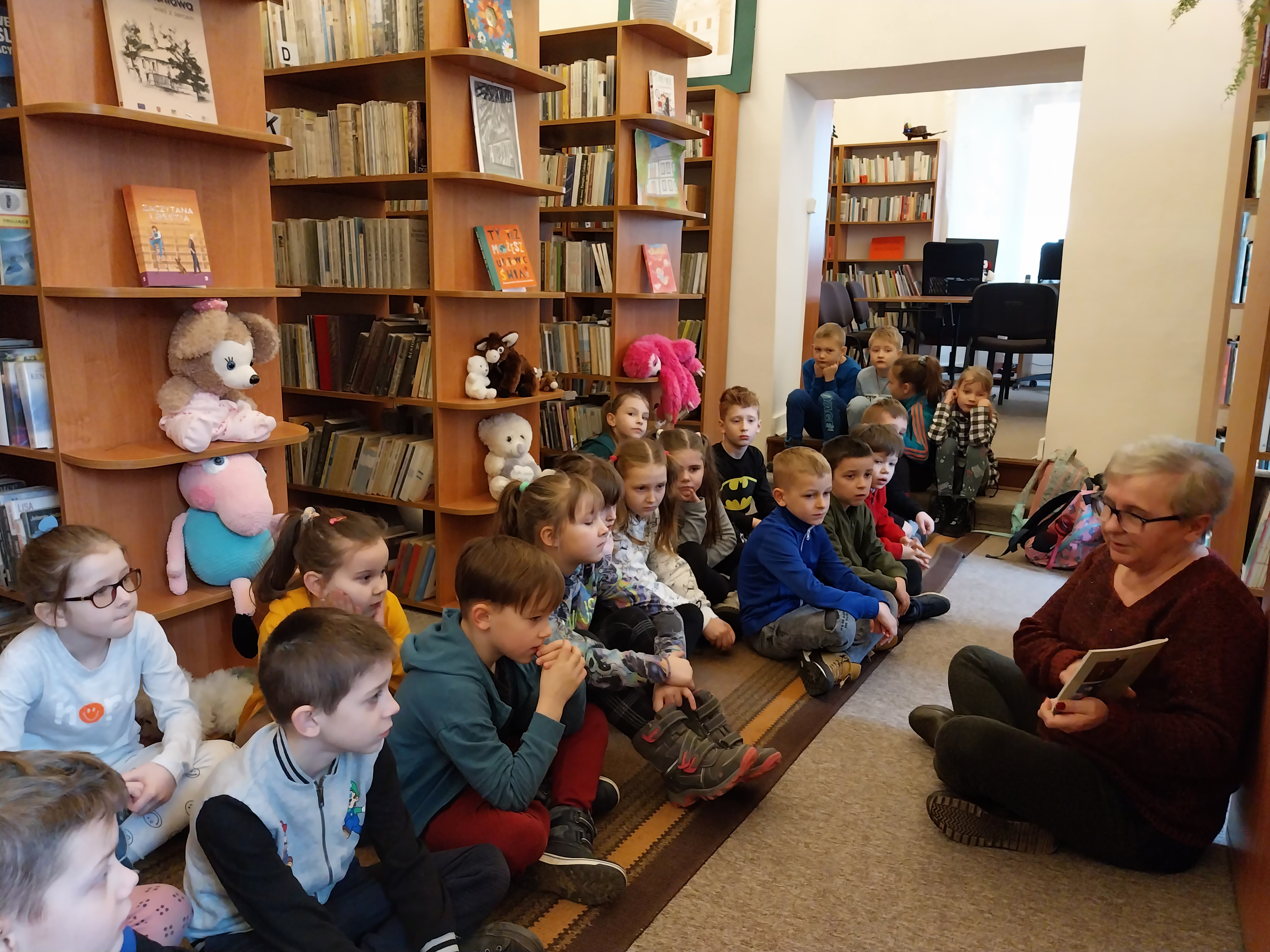 Grupa dzieci siedzi na podłodze i słucha opowiadania bibliotekarki