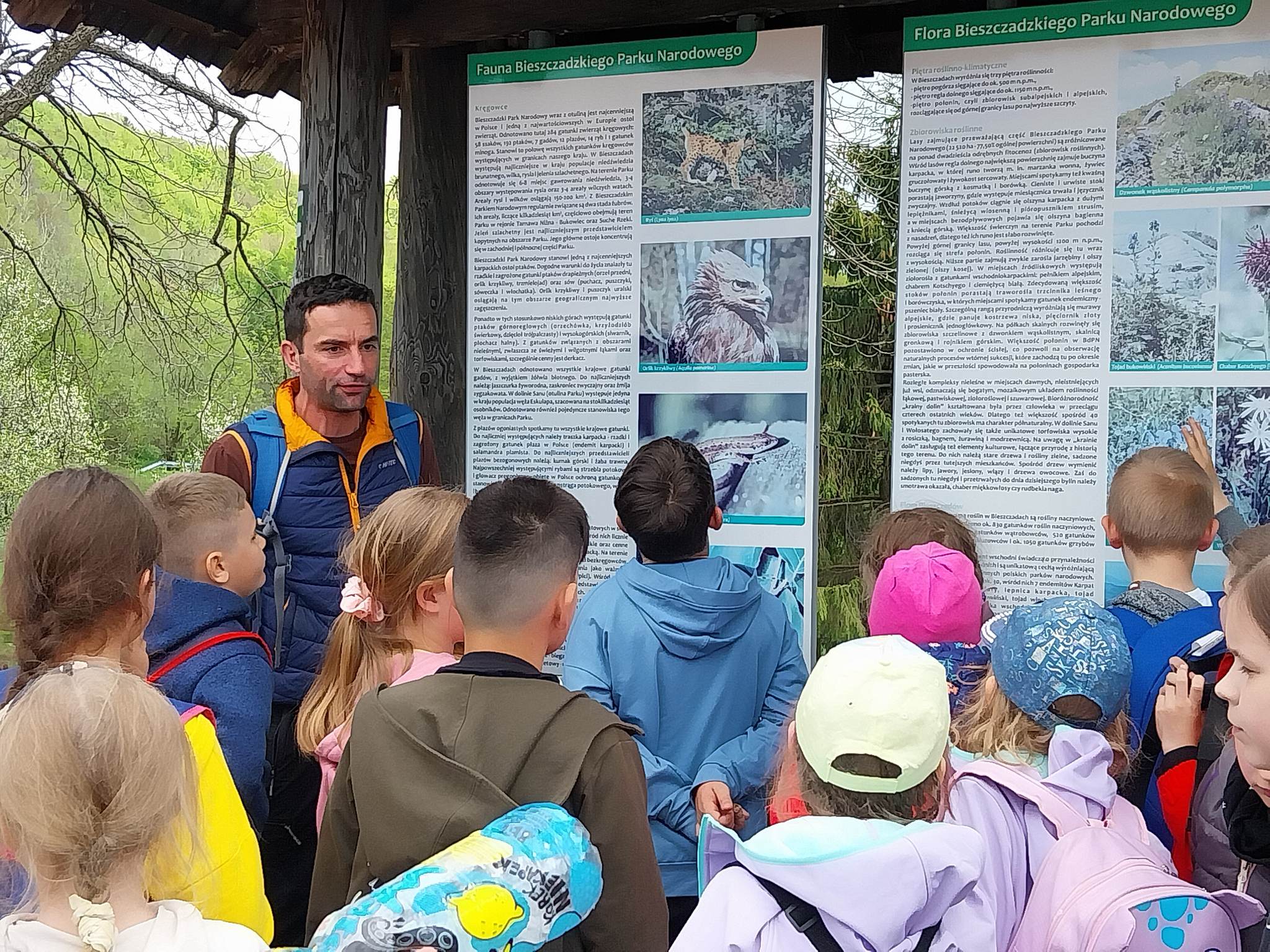 Grupa dzieci słucha opowiadania przewodnika przy tablicy informacyjnej na szlaku
