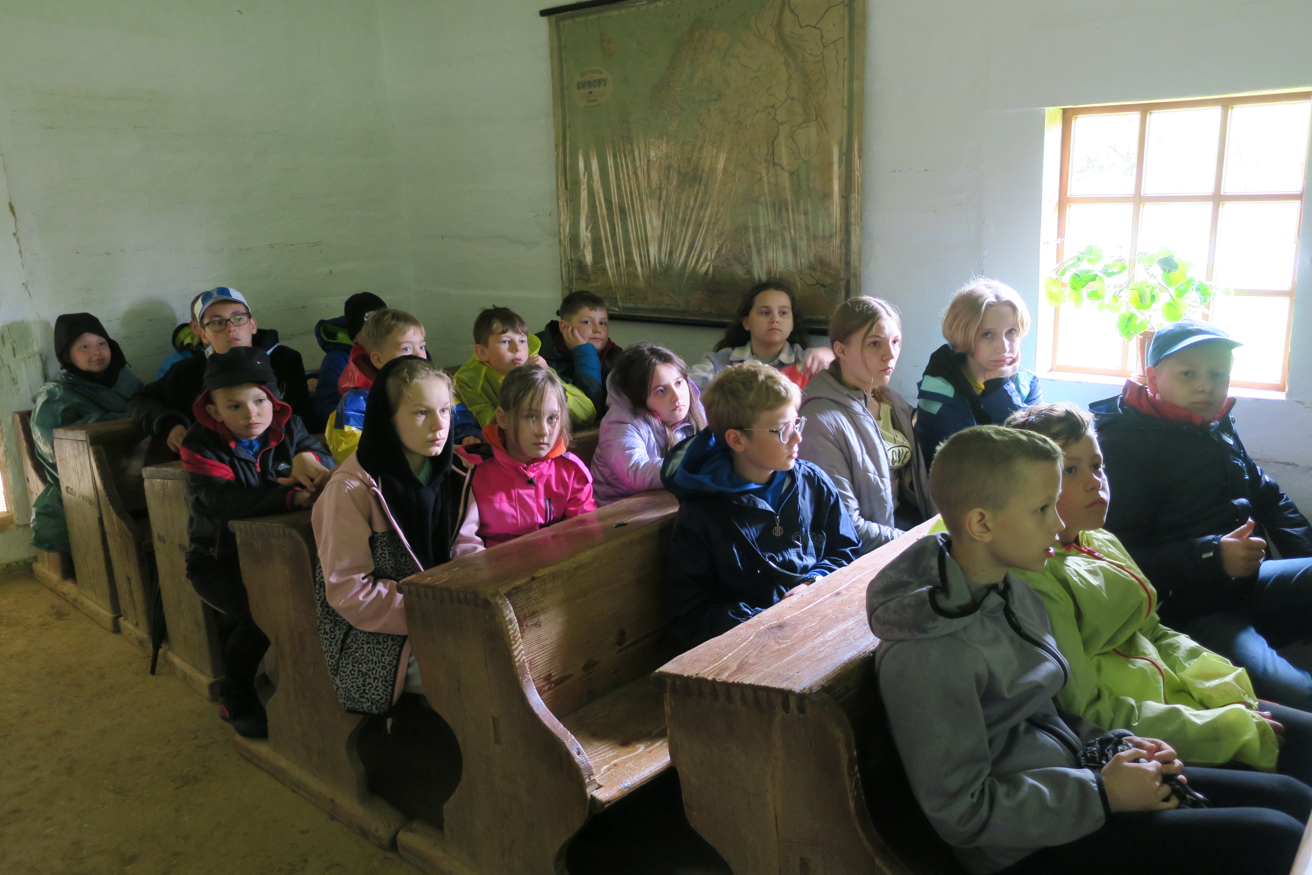 Grupa dzieci siedząca w ławkach w wiejskiej chacie