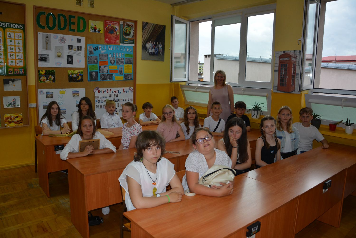Uczniowie siedzą w ławkach w sali lekcyjnej, z tyłu stoi wychowawczyni