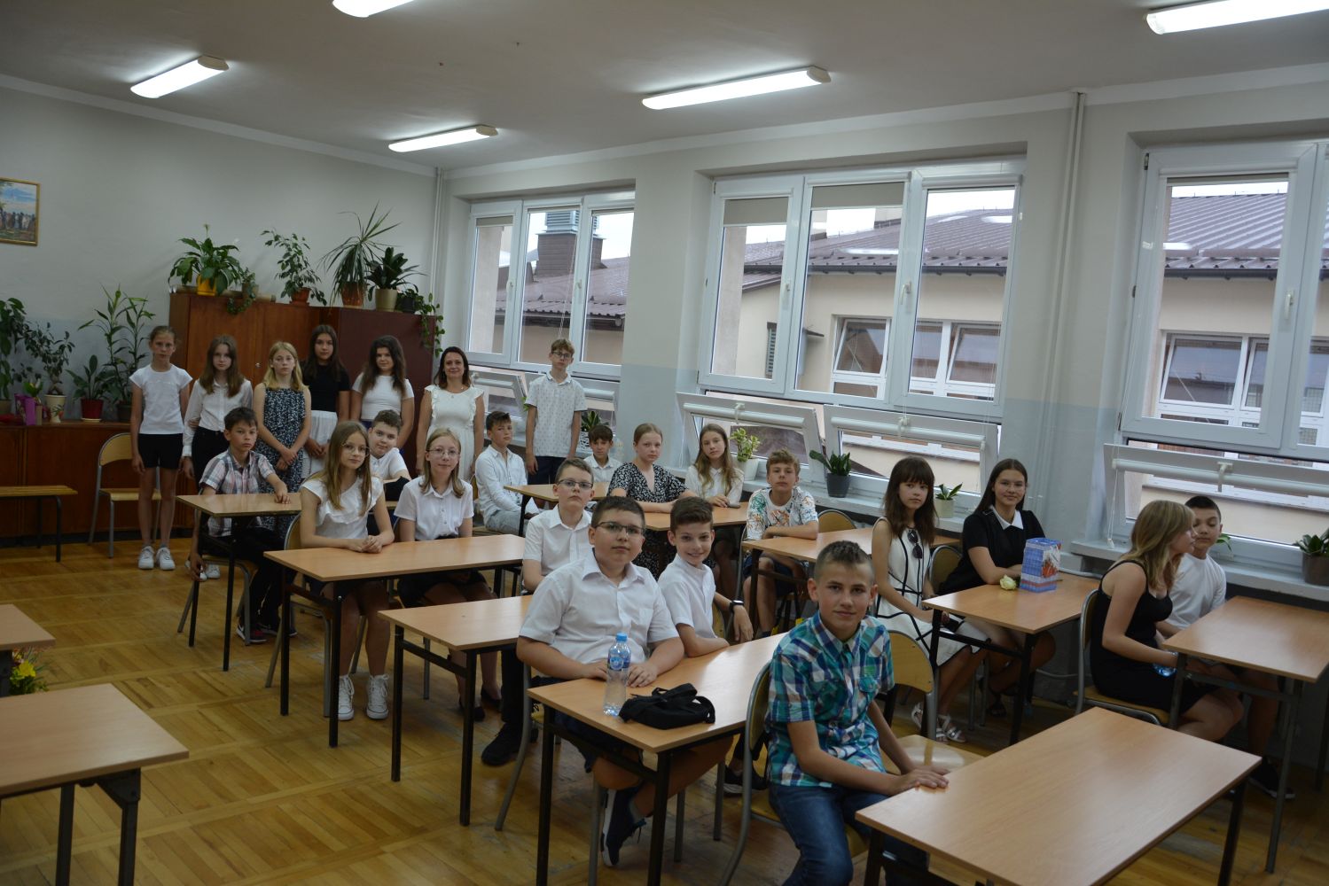 Uczniowie siedzą w ławkach w sali lekcyjnej, z tyłu stoi wychowawczyni