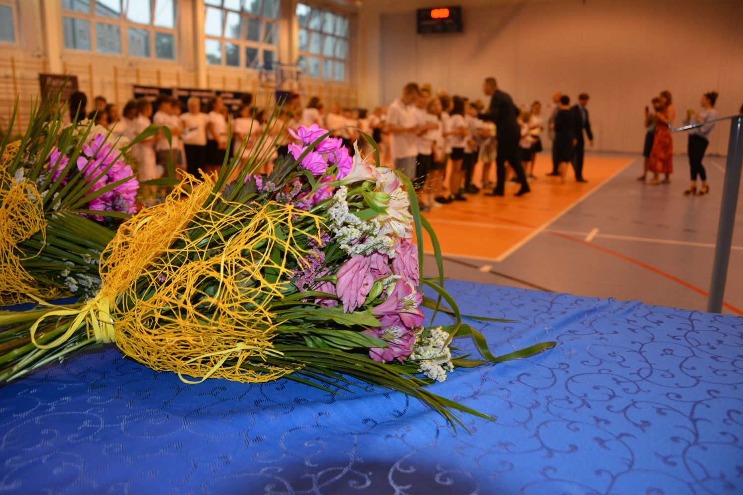 Bukiet kwiatów, w tle dyrekcja, wychowawczyni, zaproszeni goście wręczają uczniom klas trzecich nagrody książkowe