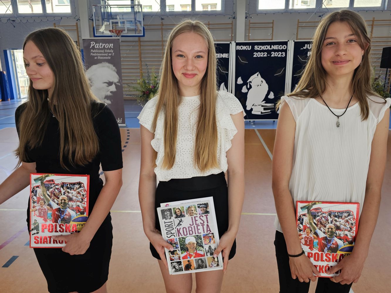 Trzy uczennice z nagrodami książkowymi za osiągnięcia sportowe stoją na tle dekoracji