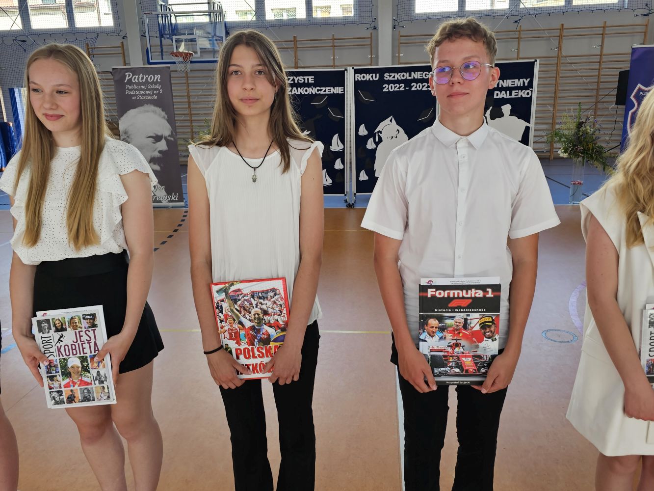 Troje uczniów z nagrodami książkowymi za osiągnięcia sportowe stoją na tle dekoracji